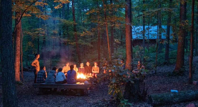 A group of people sit around a campfire in a wooded area. 